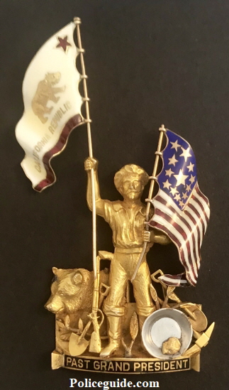 Presented to San Francisco Police Judge Matt Brady, this 1929 California NATIVE SONS OF THE GOLDEN WEST styled 14k solid gold figural badge: “GRAND PAST PRESIDENT.” These are extremely uncommon and were given out to special individuals within that group and rarely to others. The workmanship on this piece is superb with both the California Republic BEAR flag and the U.S. flag adorned with beautiful and detailed enameled work.  The theme of this piece is purely California depicting an imposing Figural GOLD MINER with his mining tools, next to an iconic California Grizzly Bear seated. Up against the miner is a gold pan with a natural native gold nugget.   The miner is gripping both flags in his two hands.  